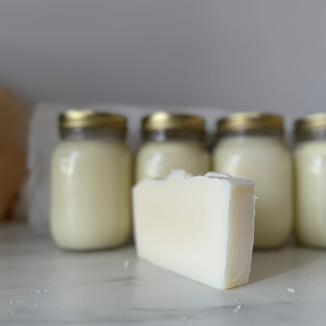 Bar of soap in front of four jars with gold lids on a light surface.