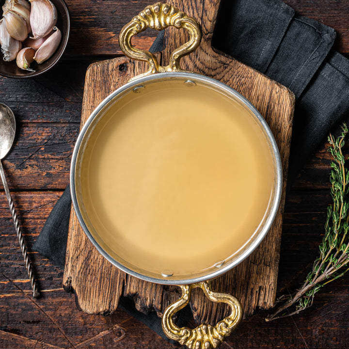 Metal bowl with bone broth on a wooden surface with a spoon and herbs.