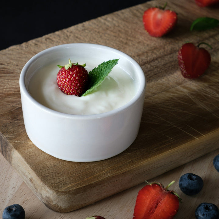 White bowl of yogurt with a strawberry on a wooden board surrounded by strawberries and blueberries.